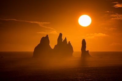 El sol de medianoche brilla sobre el mar justo al lado de la costa en la playa de arena negra de Reynisfjara.