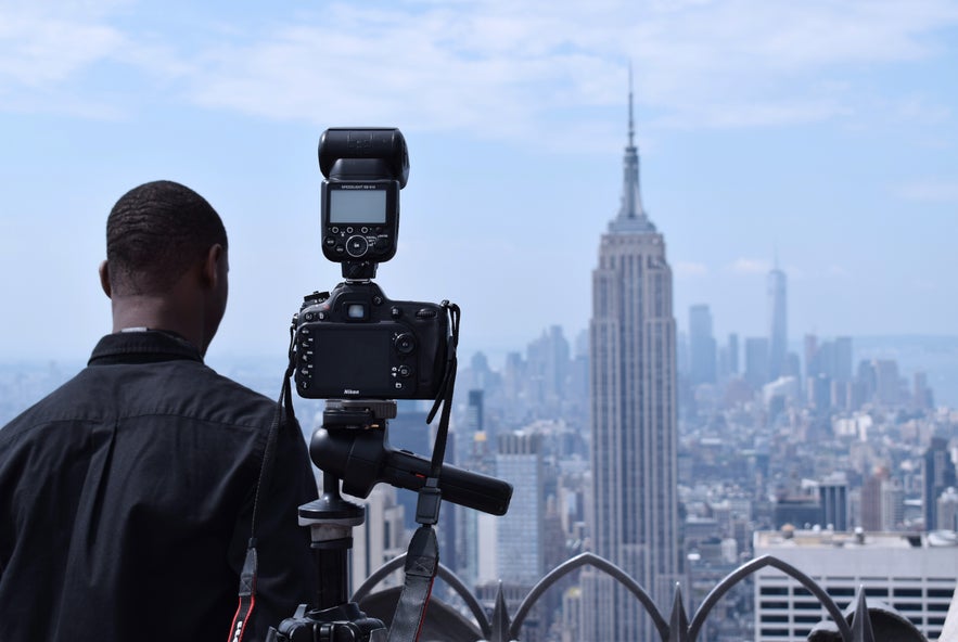 A man dressed in back has his back to the camera, standing next to a camera and flash system, pointing towards the skyline of New York City A man dressed in back has his back to the camera, standing next to a camera and flash system, pointing towards the skyline of New York City