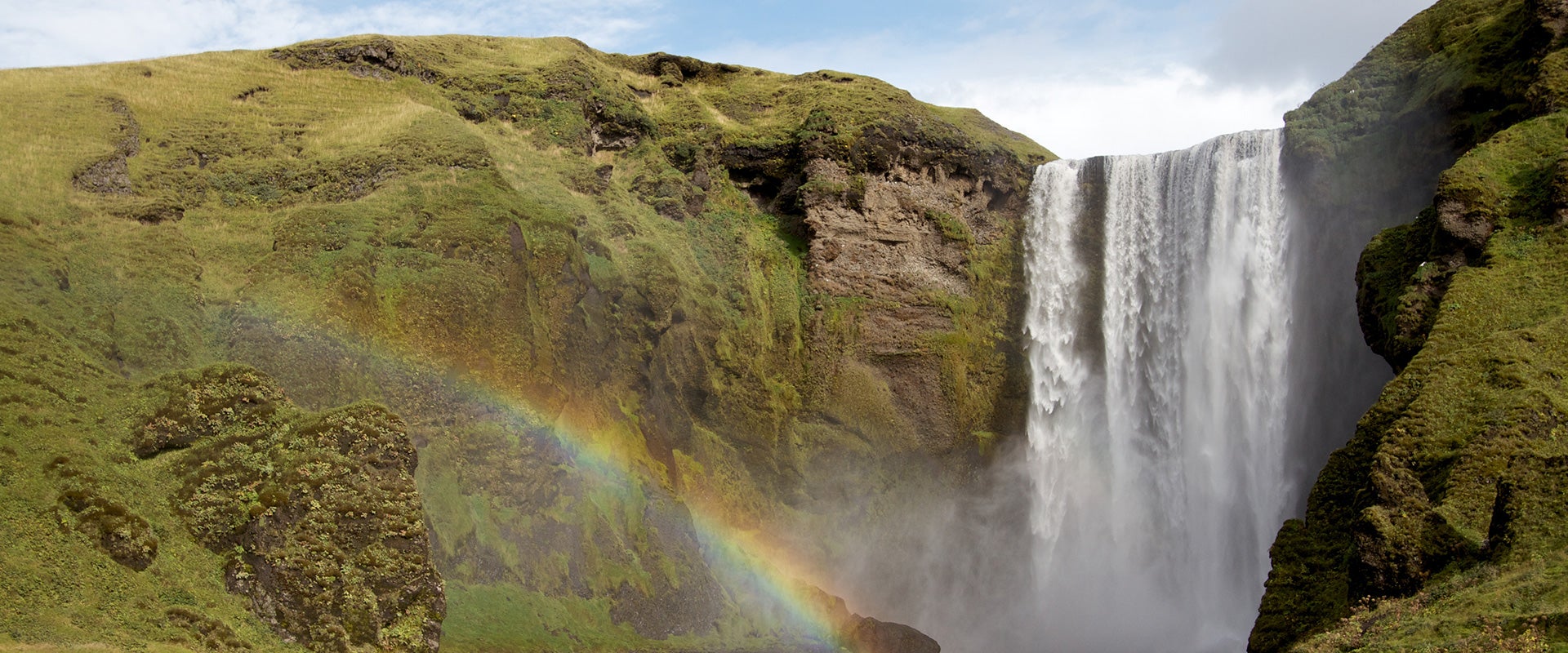 One of Iceland's stunning South Coast waterfalls features a rainbow in front on a sunny day.