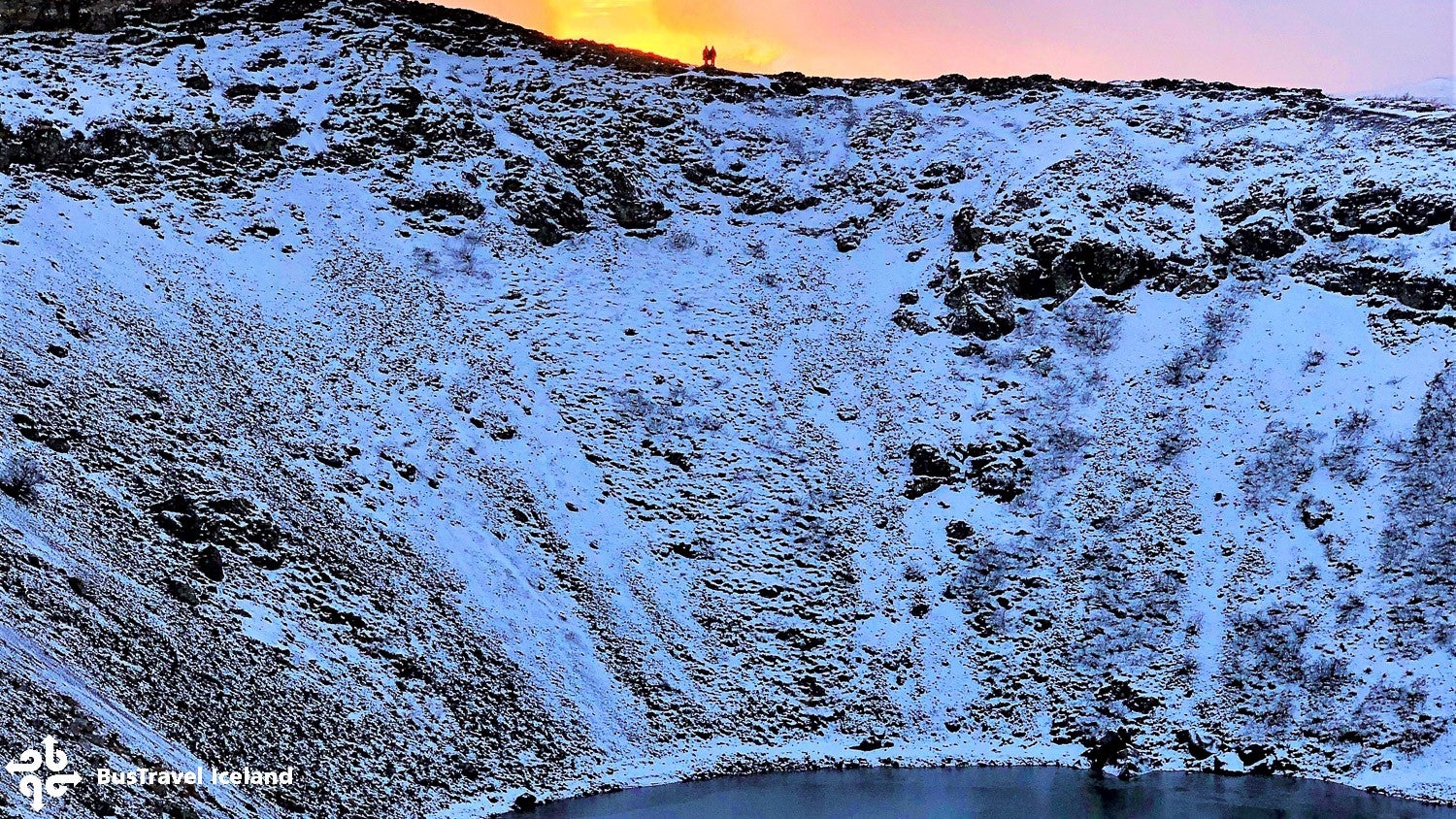 Two people stand at the top of the volcanic rock surrounding the Kerid crater during winter in Iceland.