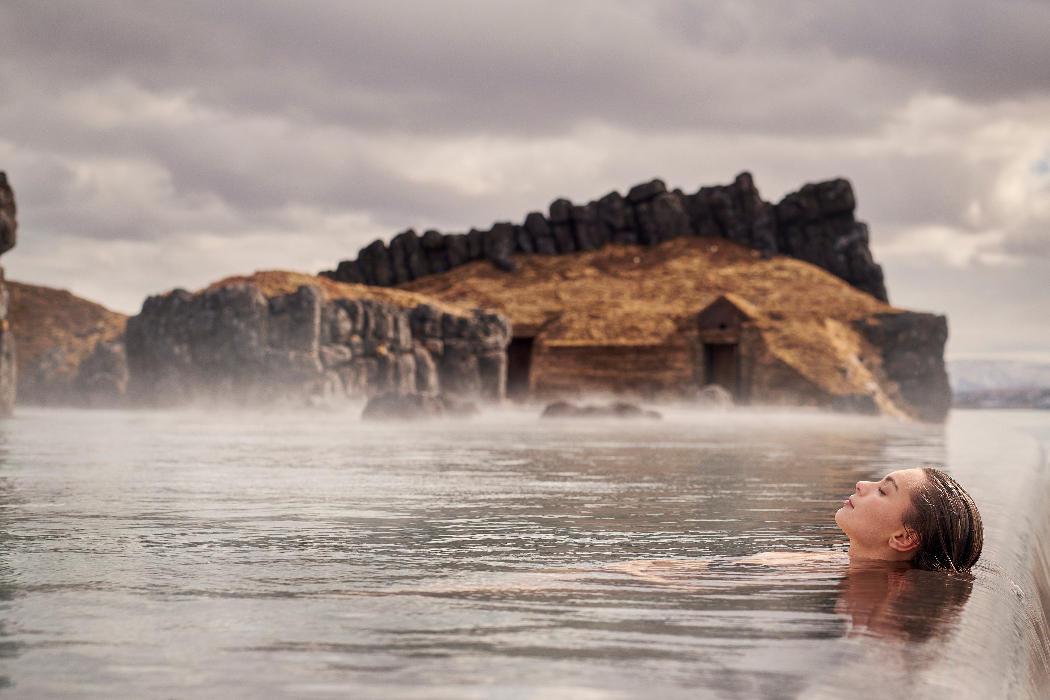 A person rests at the infinity pool edge at the Sky Lagoon spa amid a magical setting of rugged coastal scenery.