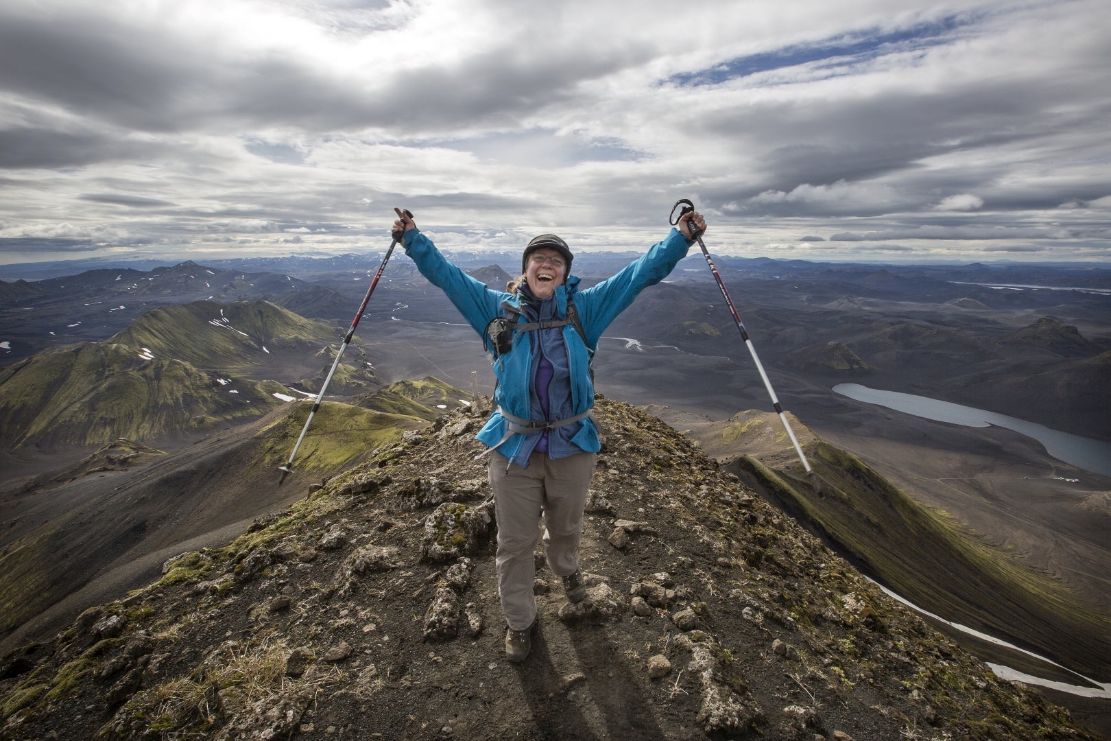 A hiker stands on a mountain in the Icelandic Highlands, raising their arms and hiking poles into the air.