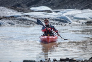 A person paddles across the calm waters of the Solheimajokull glacier lagoon with icebergs in the background.