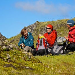 Hikers on a multi-day trek sit on a grassy hillside to enjoy their lunch on a sunny day.