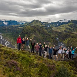 A happy hiking group stands on a ridge line in the Icelandic Highlands, with dramatic mountain and valley vistas in the background.